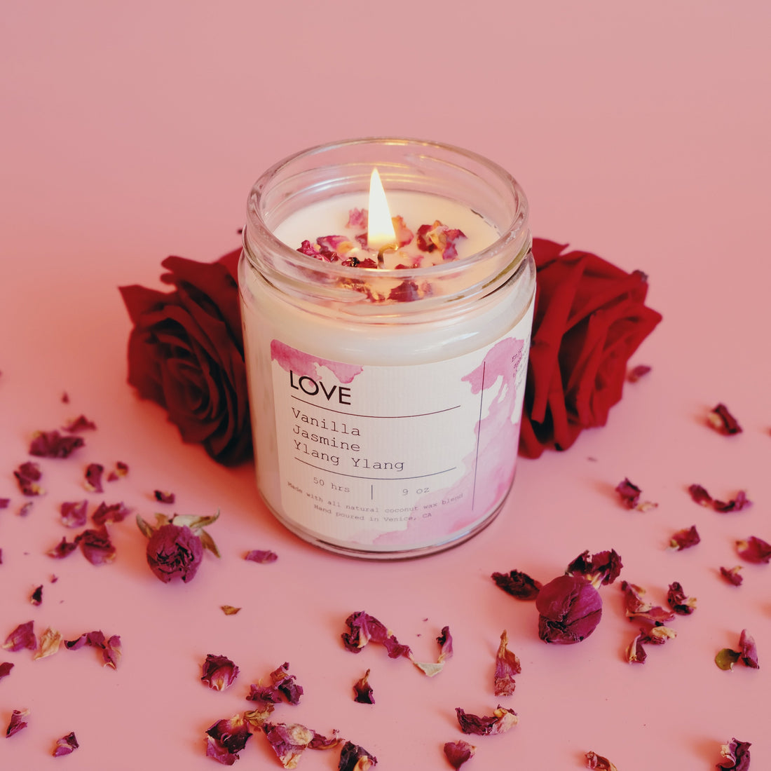 Lit candle in a glass jar with rose petals on a pink surface, flanked by red roses.