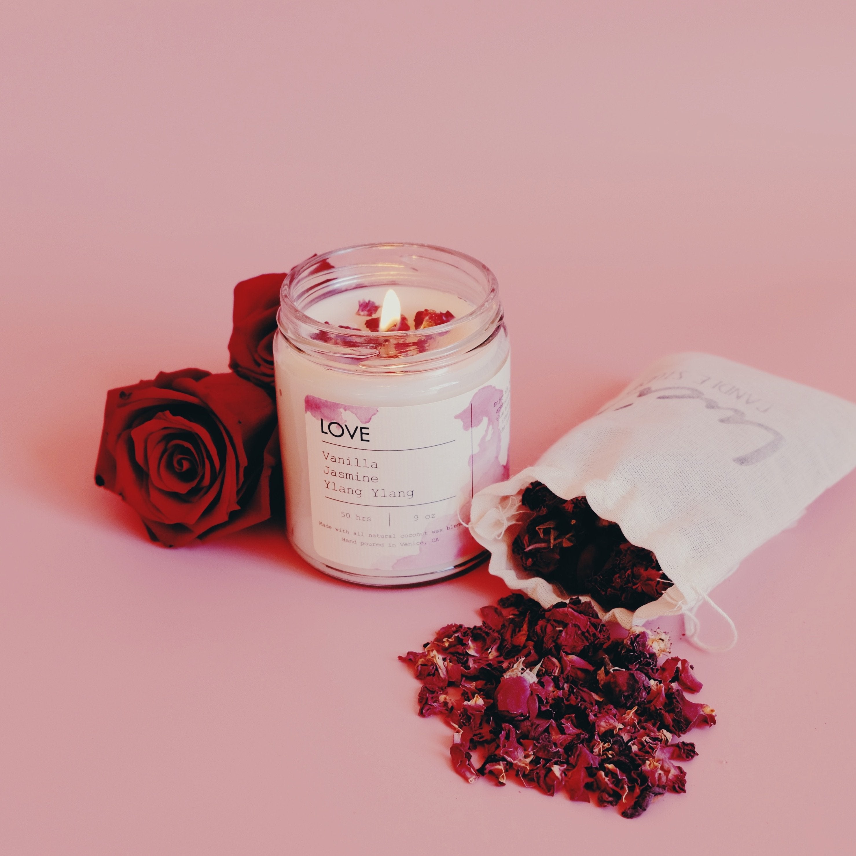 Lit candle with red roses and dried petals on a pink background, with a cloth bag beside.