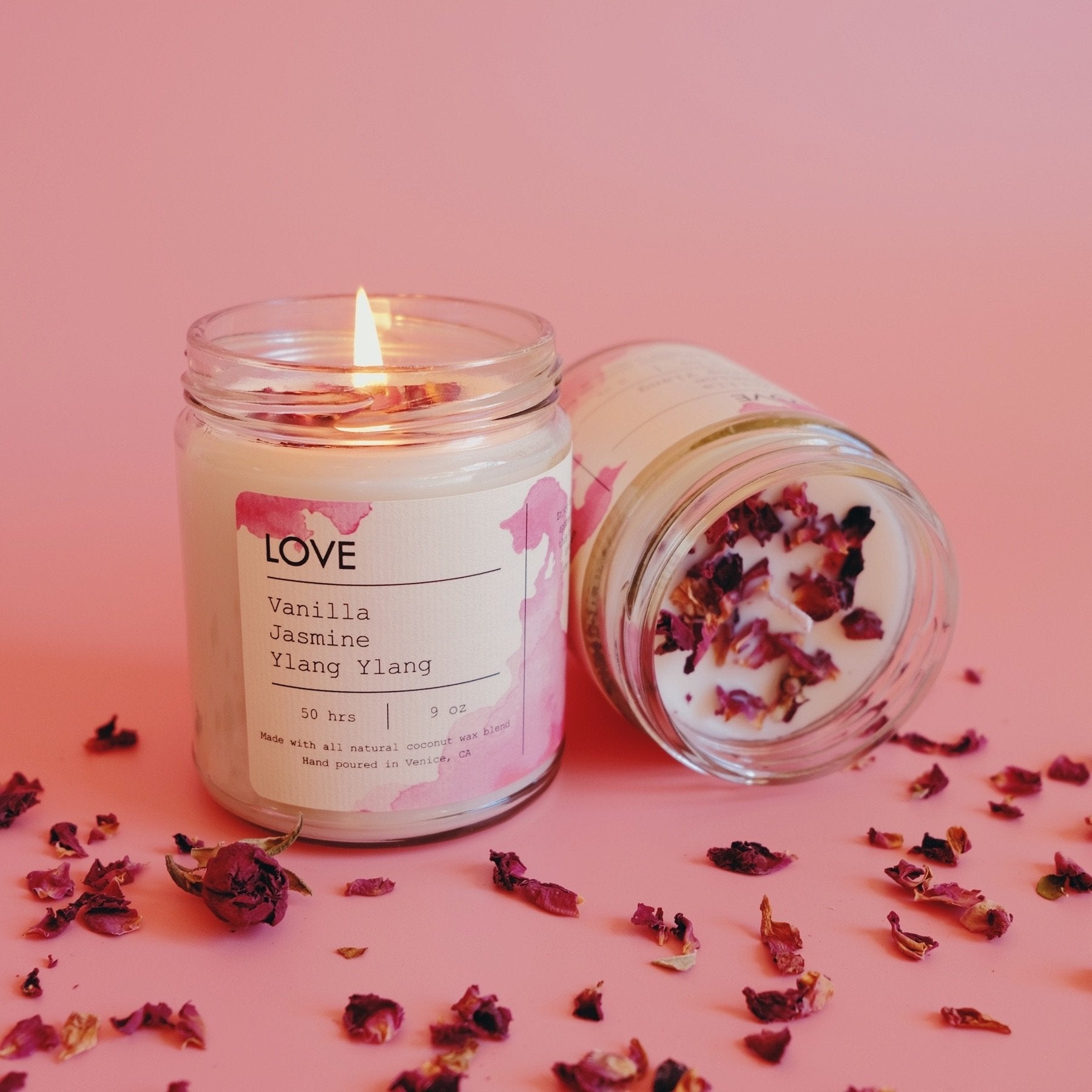 Lit candle in a jar labeled "Vanilla Jasmine Ylang Ylang," with scattered dried petals on pink.