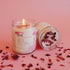 Lit candle in a jar labeled "Vanilla Jasmine Ylang Ylang," with scattered dried petals on pink.