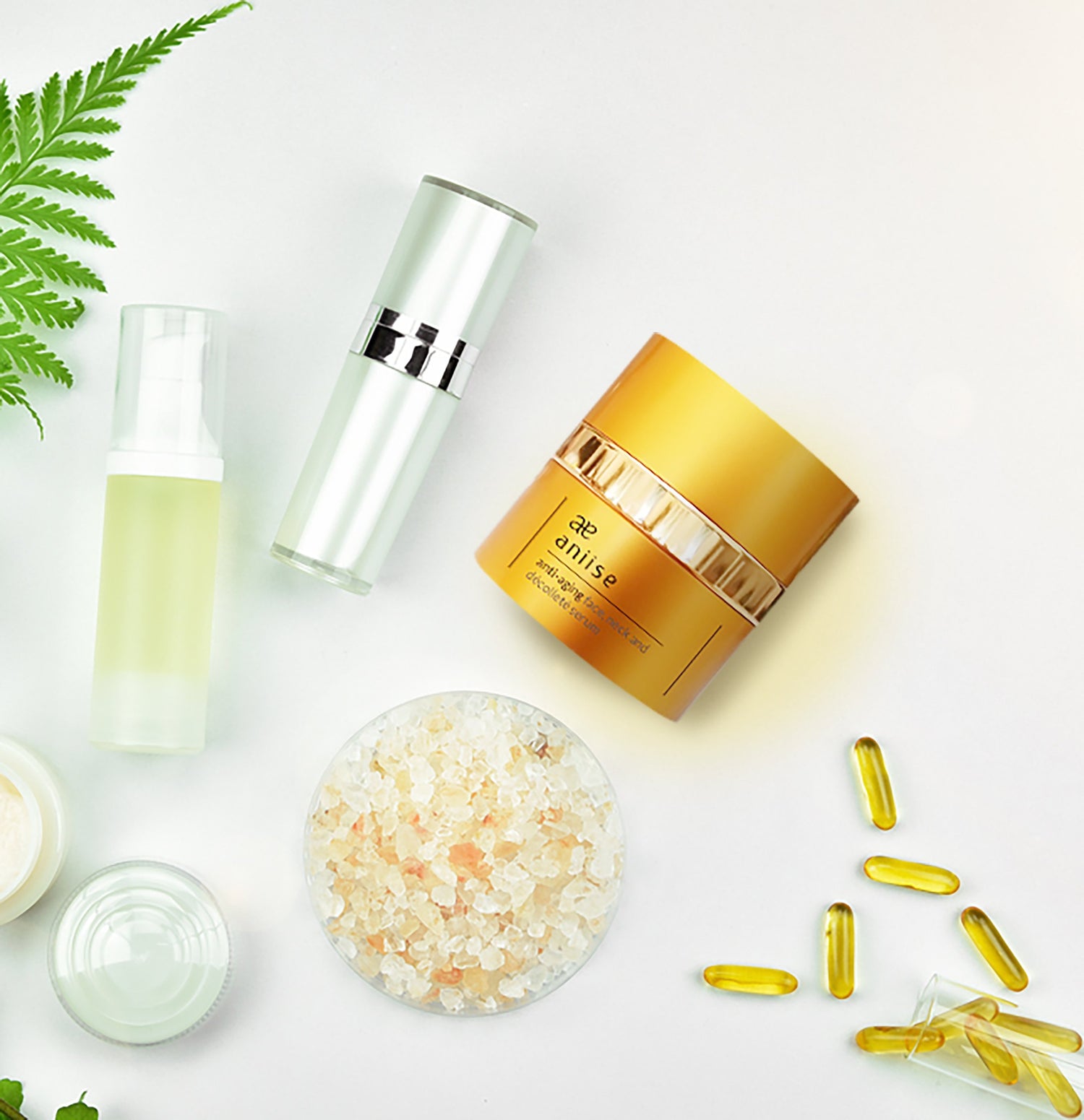 Skincare products: gold jar, silver bottles, capsules, salt, and green leaf on white background.