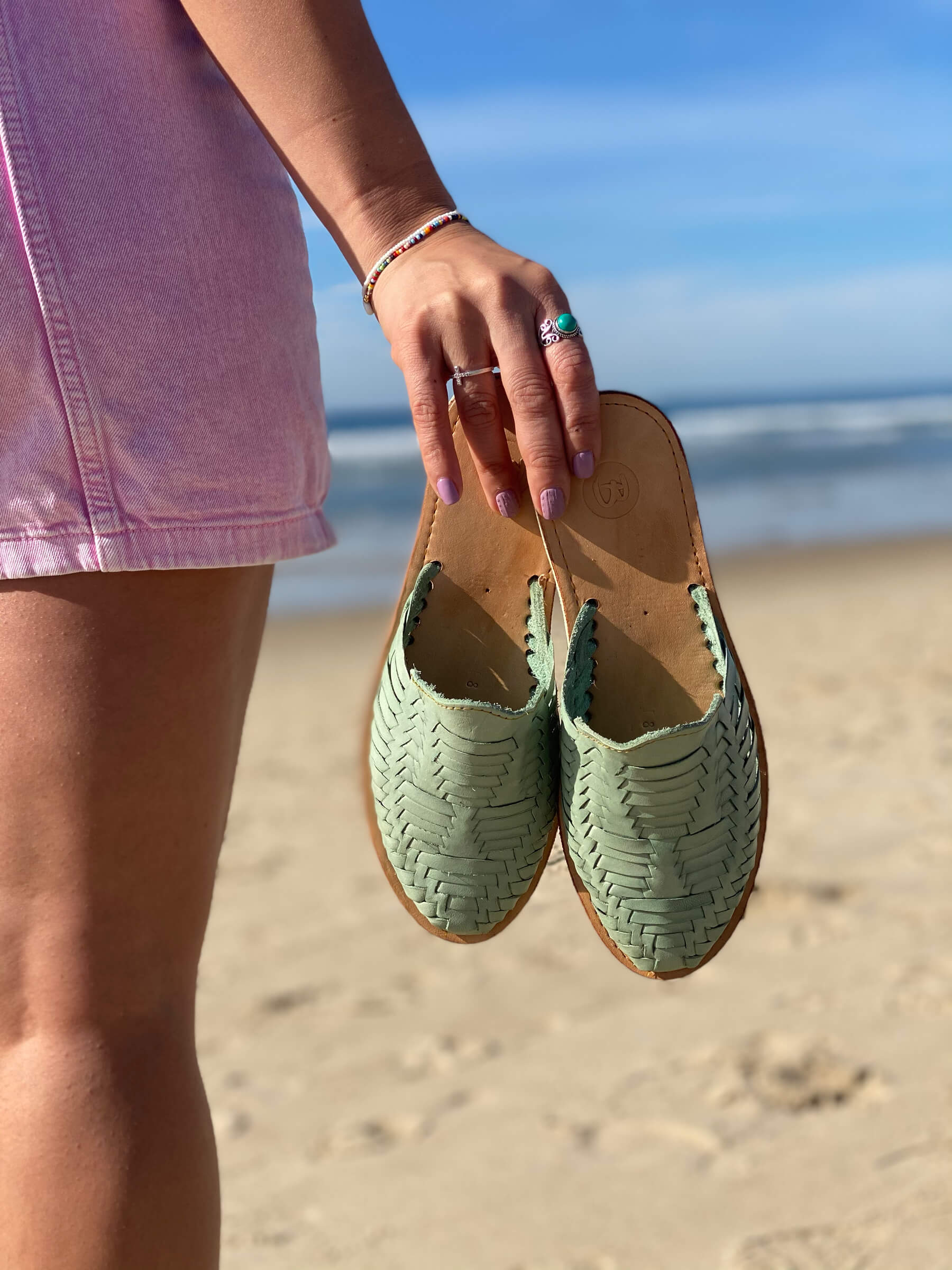 A person holds woven green shoes at the beach, wearing a pink outfit and turquoise ring.
