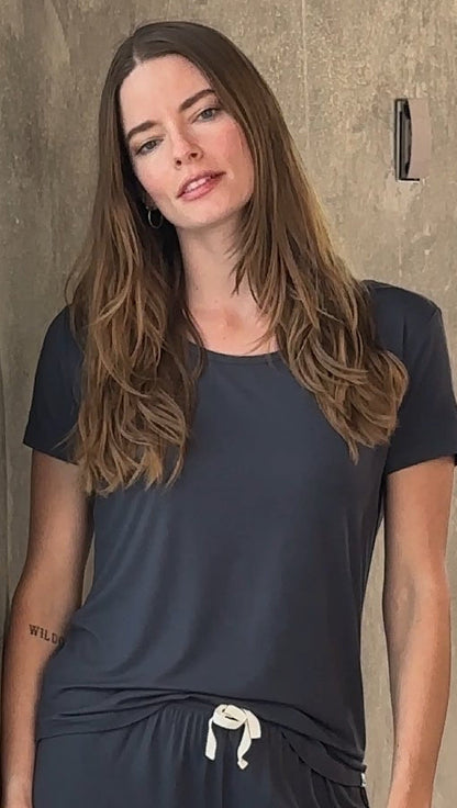 Woman with long hair in a dark gray shirt, light background, relaxed posture.