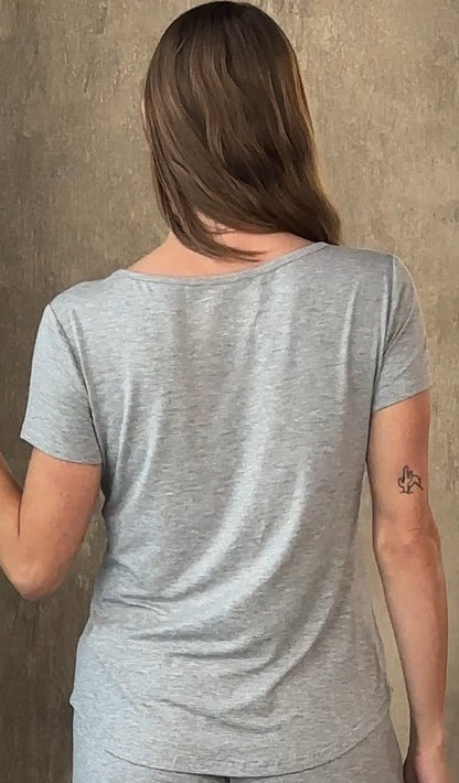 Person with long brown hair, wearing a light gray T-shirt, tattoo on arm, standing against a wall.