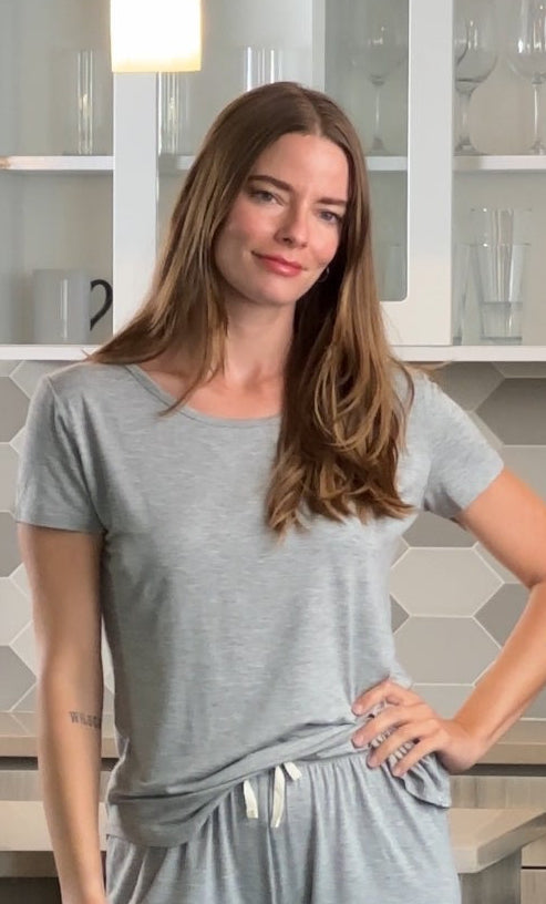 Woman with long brown hair in a gray outfit stands in a modern kitchen with hexagonal tiles.