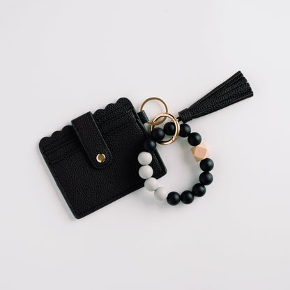Black cardholder with scalloped edge, tassel keychain, and black and white beaded bracelet.