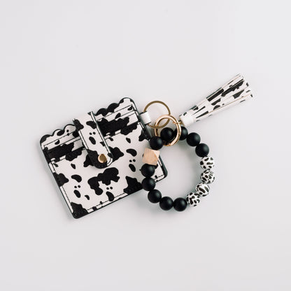 Cow-print cardholder with scalloped edges, gold button, and black bead bracelet with tassel.