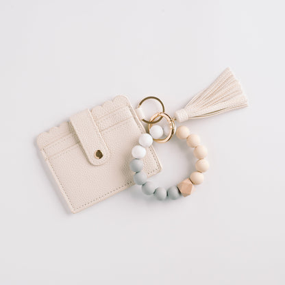Cream cardholder with scalloped edges, tassel, and a beaded key ring in neutral tones.