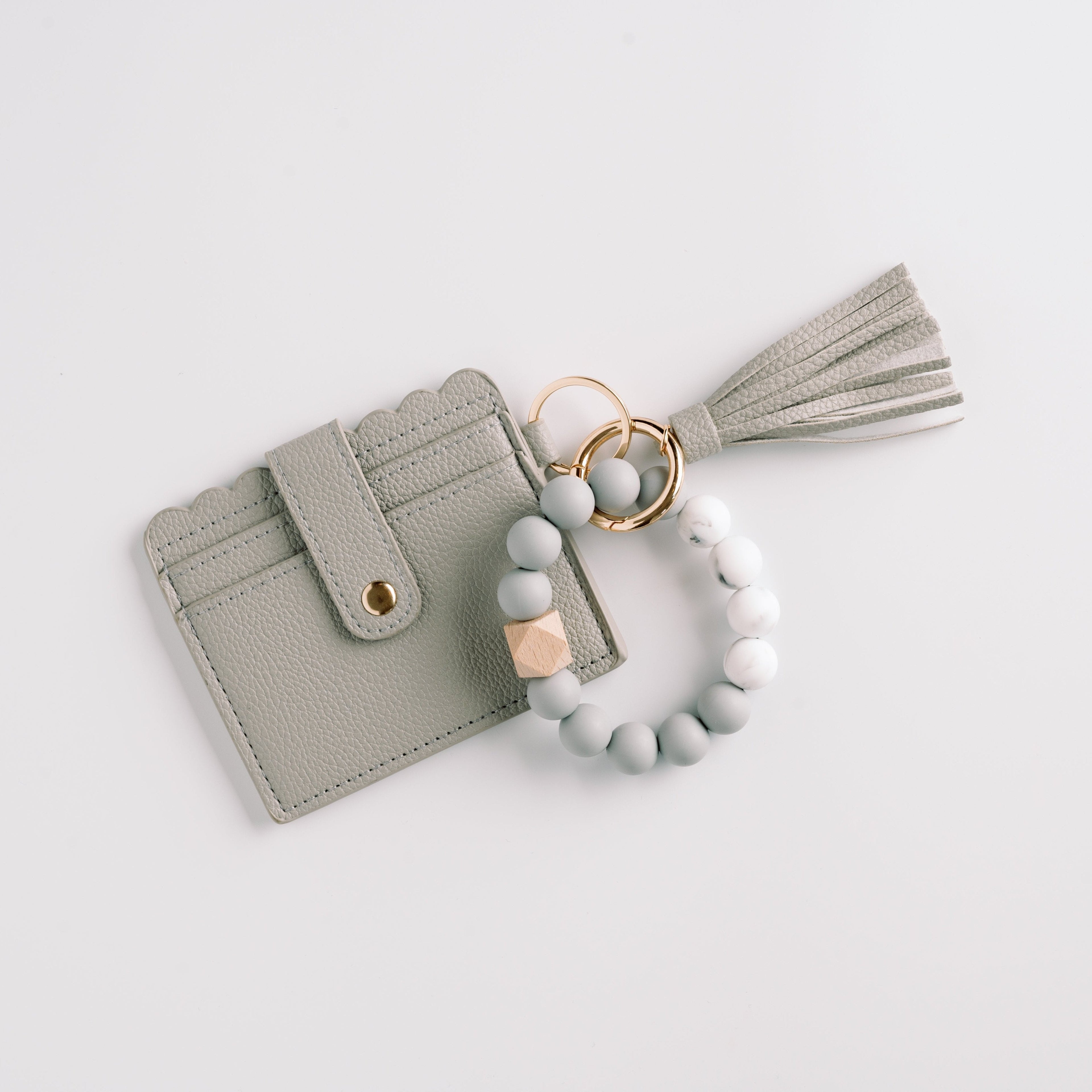 Gray cardholder with scalloped edges, gold button, tassel, and beaded wristlet on white background.