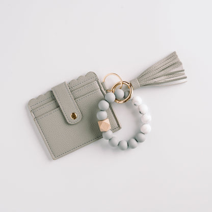 Gray cardholder with scalloped edges, gold button, tassel, and beaded wristlet on white background.