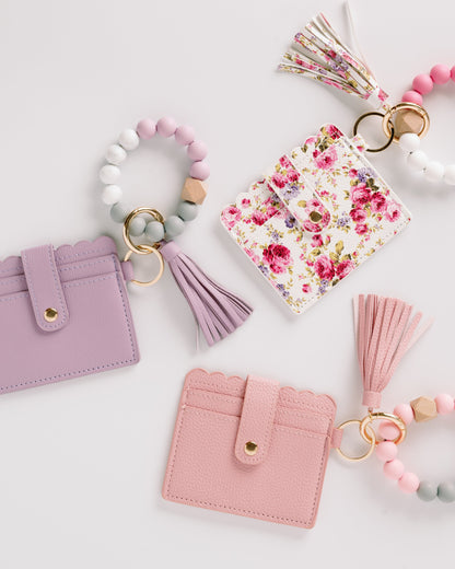 Three cardholders in pink, purple, and floral designs, each with a matching tassel and bead keychain.