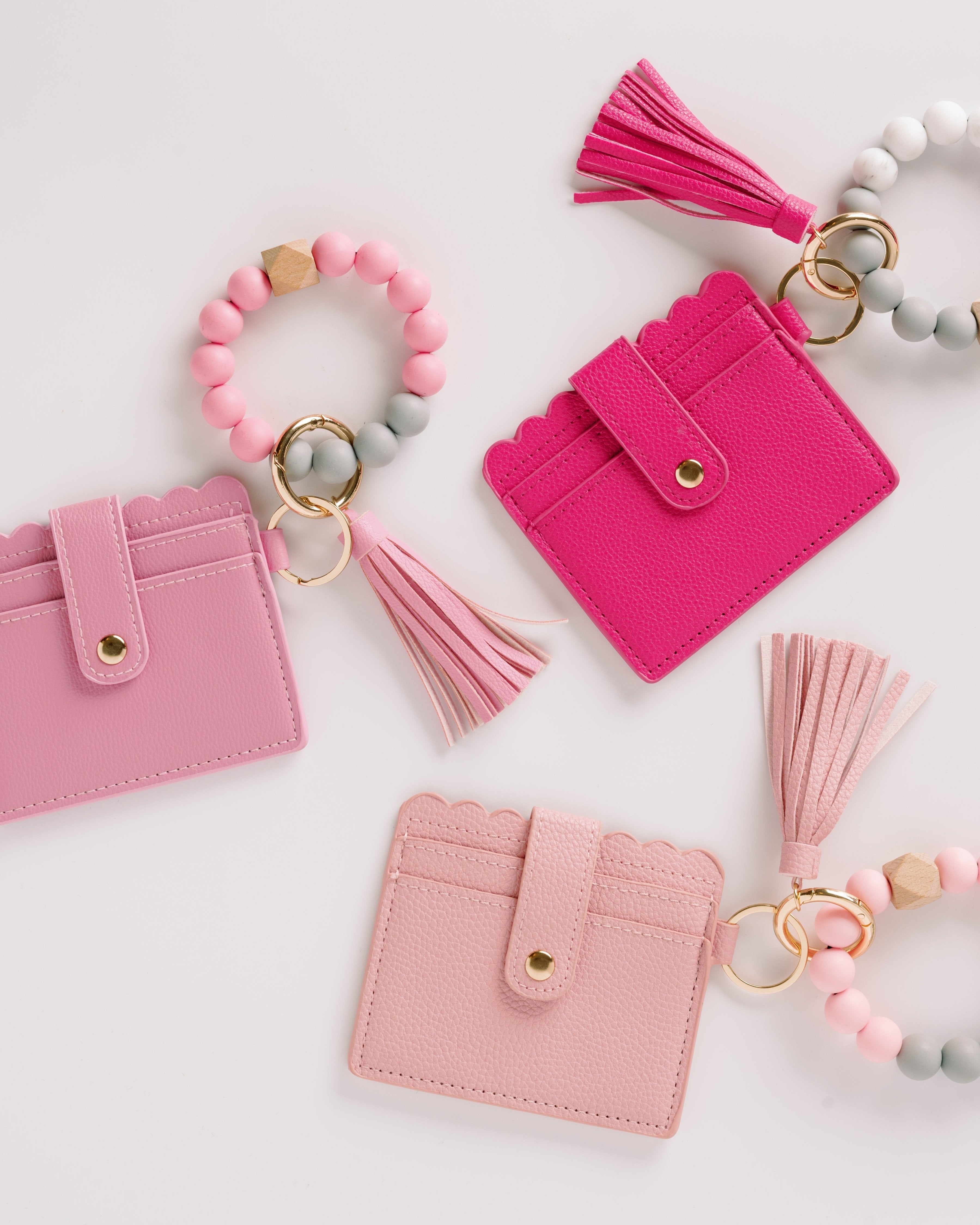 Pink and blush scalloped wallets with tassels and beaded keychains on a white background.