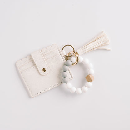 White cardholder with scalloped edges, beaded keychain, gold rings, and tassel on white background.