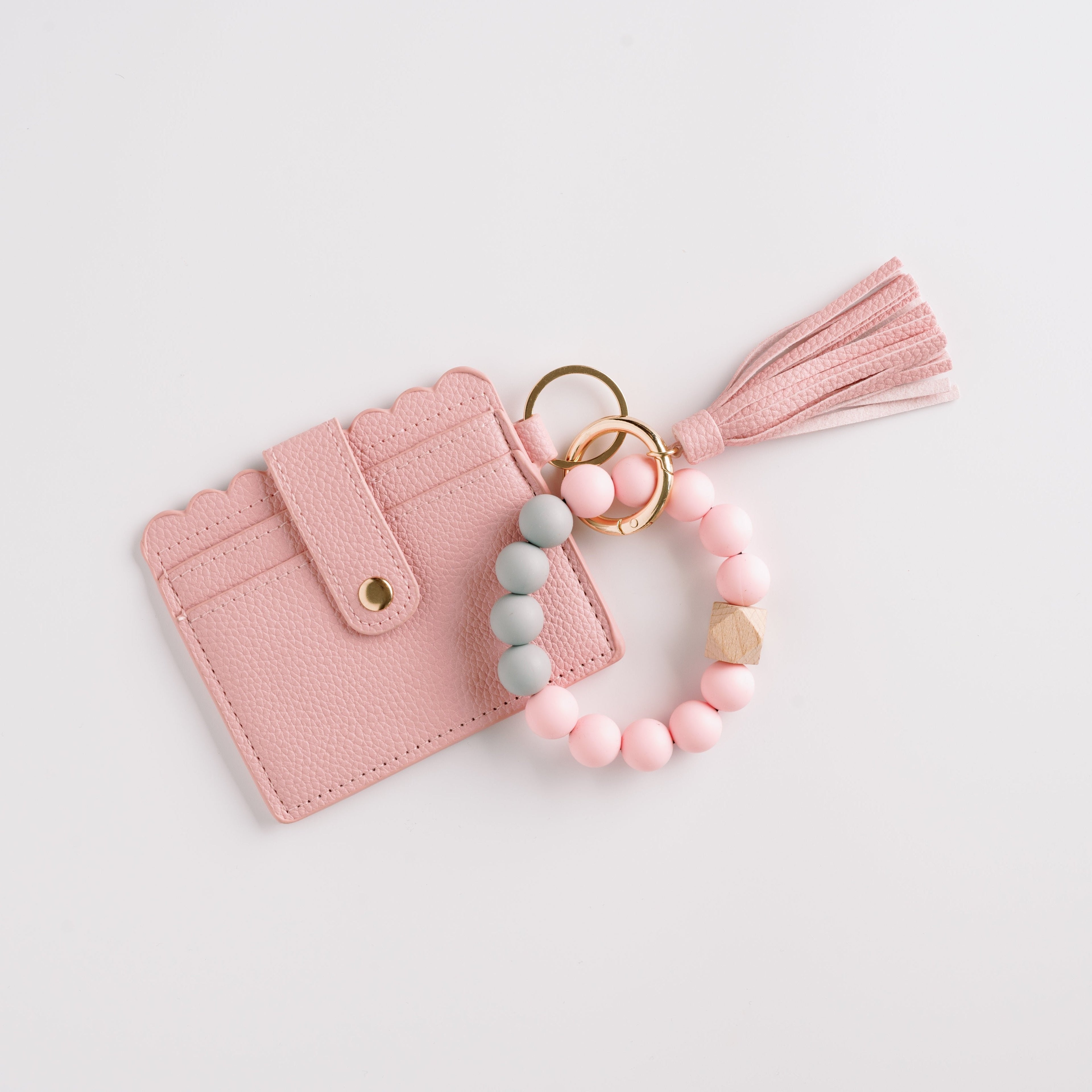 Pink scalloped wallet with tassel, linked to pastel bead bracelet and gold rings on white background.