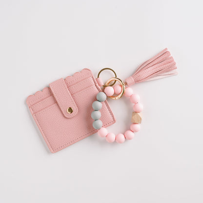 Pink scalloped wallet with tassel, linked to pastel bead bracelet and gold rings on white background.