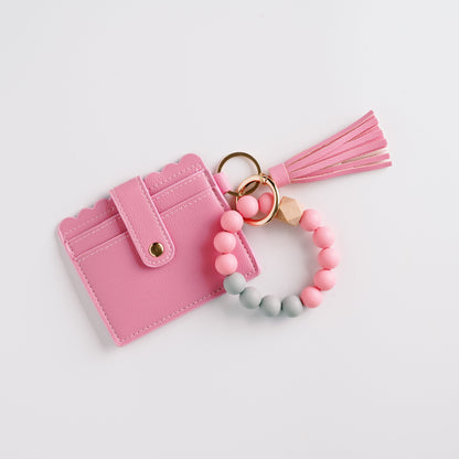 Pink cardholder with a tassel, paired with a pink and gray beaded bracelet on white background.