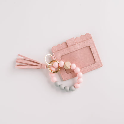 Pink cardholder with a tassel, linked to a keyring of pastel pink and gray beads on white.