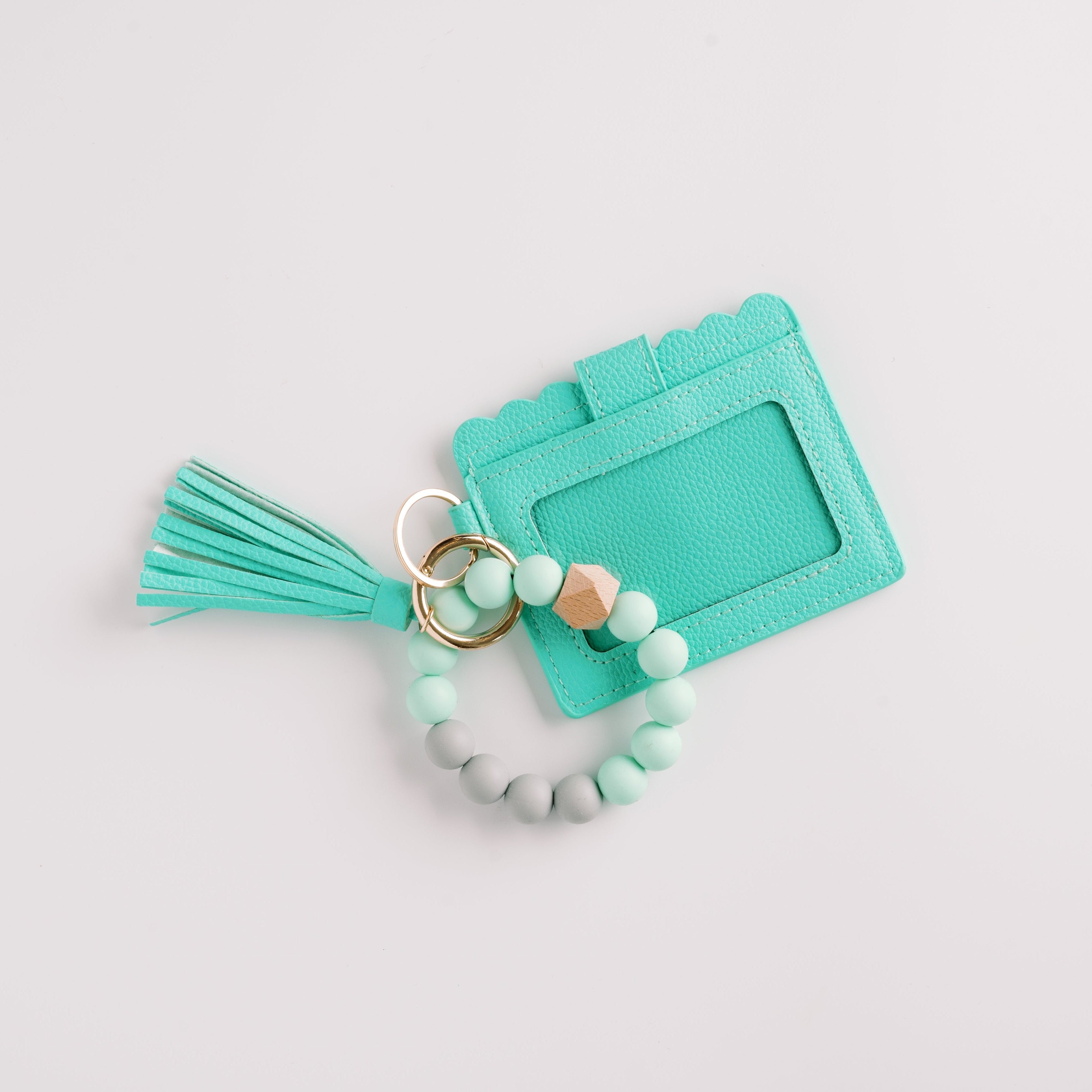 Mint cardholder with scalloped edges, tassel, bead bracelet, and gold rings on a white background.